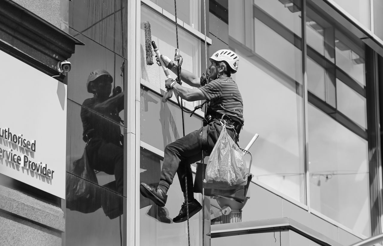 hero-img Black and white from below of man steeplejack in protective helmet with professional equipment cleaning glass wall of modern tower